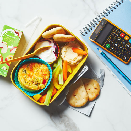 Metal bento box filled with frittata cups, hummus and mini pita breads, and a berry bottom yogurt with a notebook sitting next to it