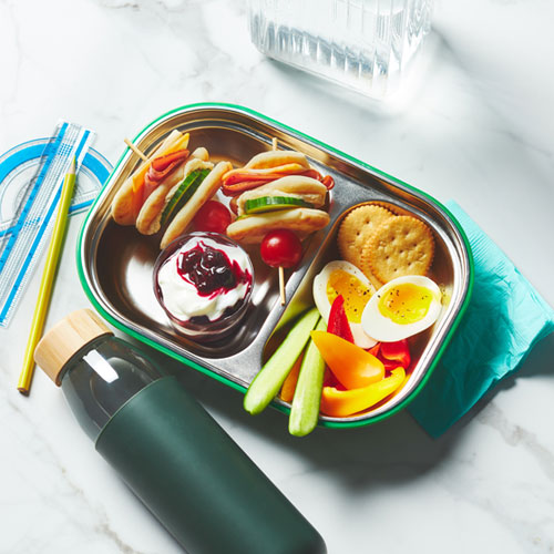 Metal bento box with ham and hummus skewers, mixed veggie and cracker snacks and a layered berry parfait sitting next to a green water bottle.