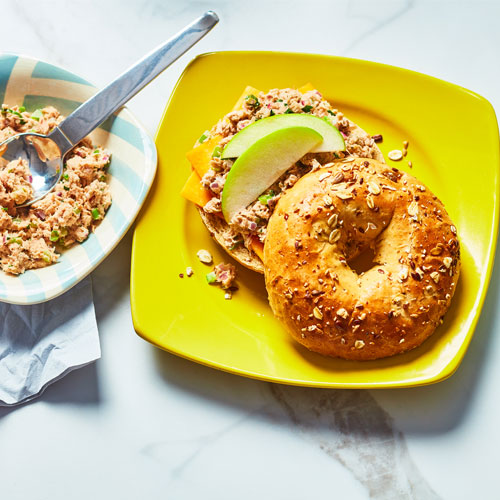 Multigrain bagel topped with a tuna salad, melted cheese and apple slices on a bright yellow plate.