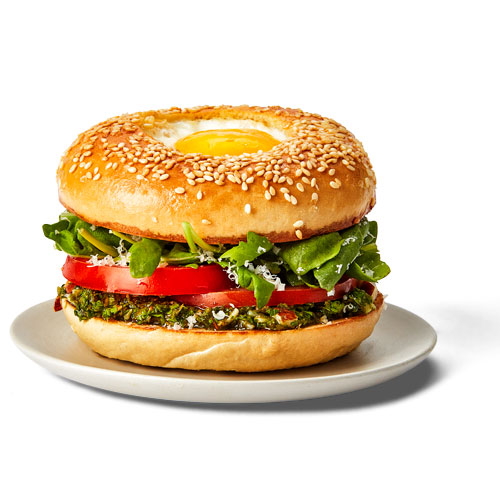 Sesame bagel topped with basil pesto, fried egg, tomato slices, Parmesan cheese and baby arugula on a white plate.