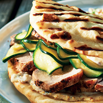 Grilled naan bread sandwiching grilled pork medallions and quick zucchini pickles, sitting on a white plate next to a side of coleslaw.