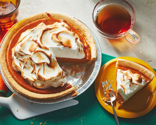 Pumpkin pie with clouds of meringue on top sitting next to a slice of the pie on a yellow plate. 