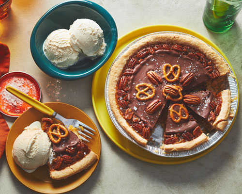 Pecan pie topped with chocolate ganache, pecans and sea salt flakes with a bowl of ice cream in a dish next to it and a slice on a yellow plate. 