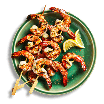  A round green plate with three cooked shrimp skewers laid on top and cut limes for garnish.