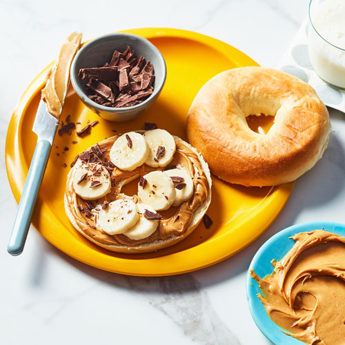 Open faced plain bagel topped with peanut butter, sliced bananas and chocolate shavings on a bright yellow plate. 