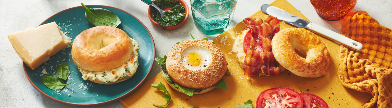 Three different bagels with different toppings including eggs and bacon, sit atop a mustar yellow board and a bule plate.