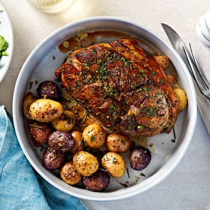 Boneless leg of lamb roasted in a white baking dish alongside roasted new potatoes on a white countertop, next to a fresh green salad.