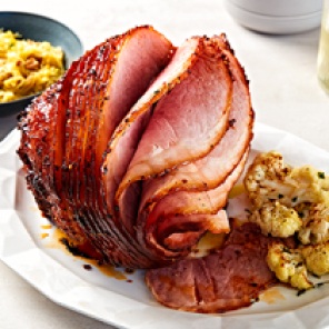 Glazed, sliced Panache hickory smoked honey spiral ham on a white platter with roasted cauliflower on the side, next to a carving fork and knife.