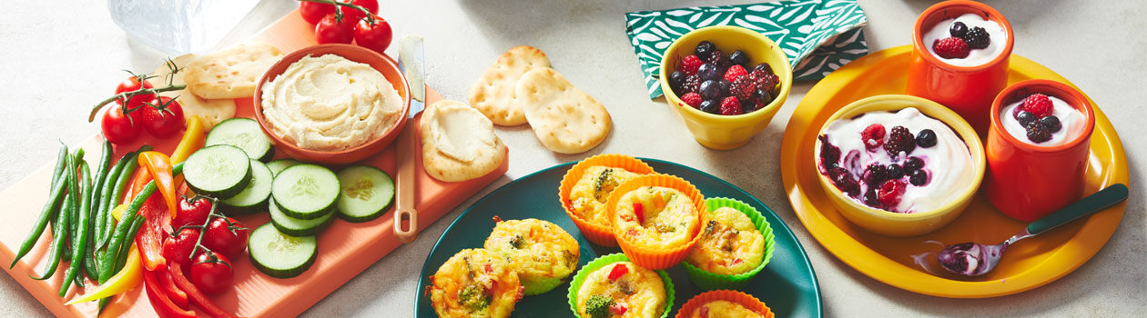 Variety of kid-friendly lunches including dips and veggies, mini frittatas and yogurt parfaits on colourful boards and plates.