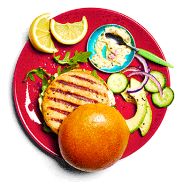 Cooked and dressed wild sockeye salmon burger on a red plate next to sliced avocado and onion.