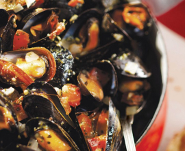 A close up of opened mussels in a bowl flecked with pesto, onions and tomatoes.