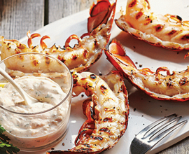 Grilled lobster tails on a platter beside a cup of aioli with porcelain spoon beside two lemon wedges.