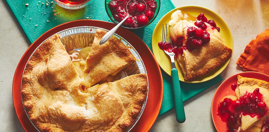 Baked apple pie sitting next to a slice with a spiced cranberry sauce drizzled on top. 