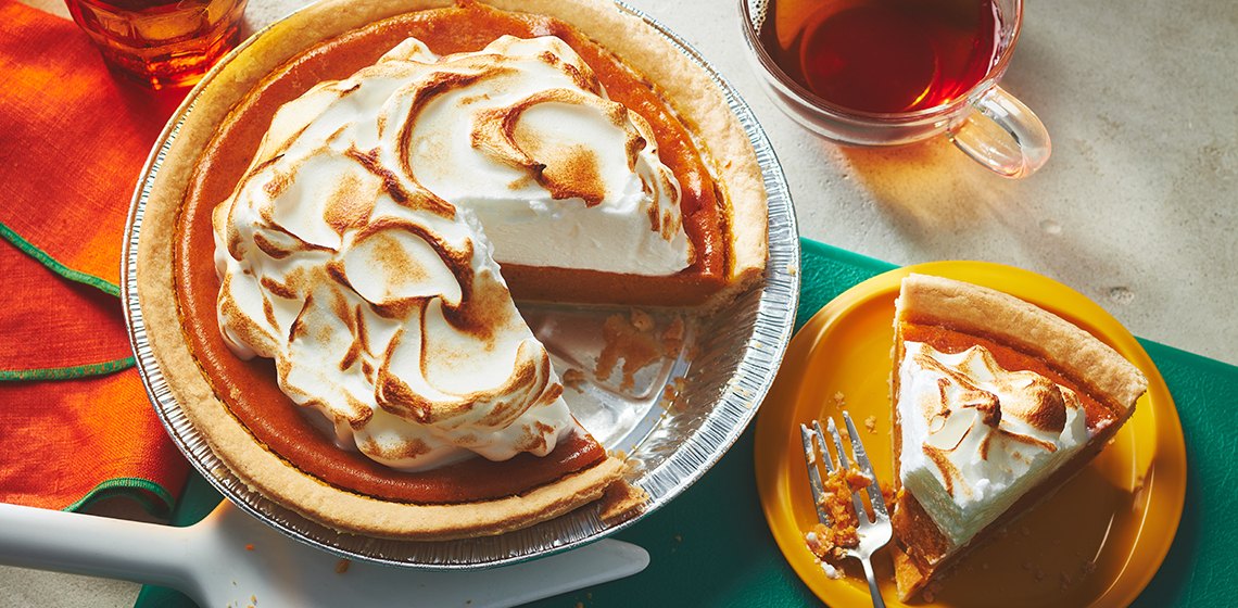 Pumpkin pie with clouds of meringue on top sitting next to a slice of the pie on a yellow plate. 