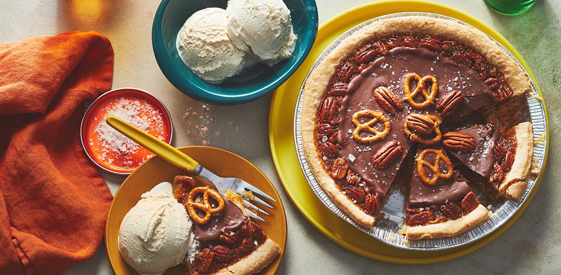 Pecan pie topped with chocolate ganache, pecans and sea salt flakes with a bowl of ice cream in a dish next to it and a slice on a yellow plate. 