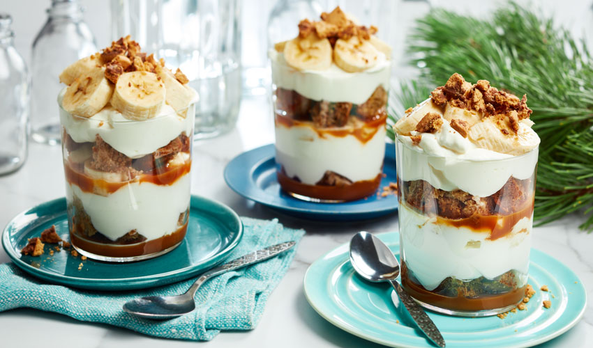 Individual serving glasses filled with Banoffee trifle, sliced bananas, whipped cream, chocolate coated digestive cookies and dulce de leche spread.