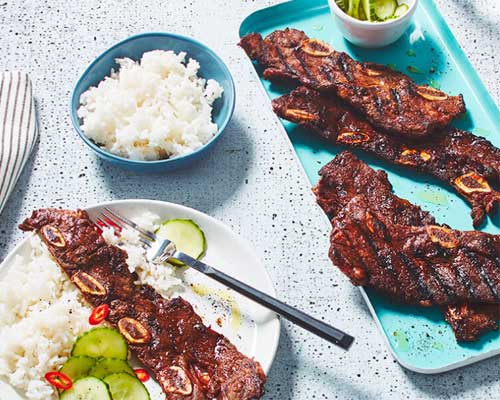 white plate with fork, rice pilaf, and cucumber salad beside blue bowl of rice and sperate blue serving dish of short ribs.