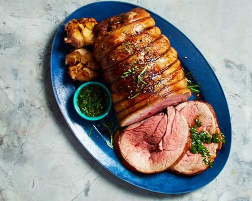 Top-down view of a place setting with circular dish of lamb and roasted garlic on a blue plate.
