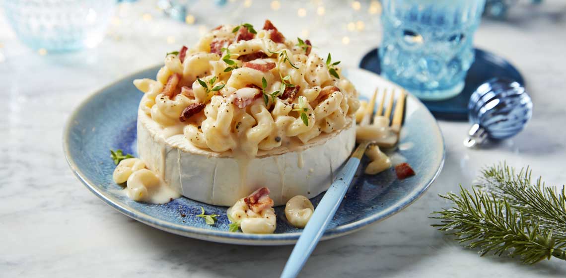 Mac n’ cheese mounded on top of a hollowed out brie wheel on a blue plate with a fork sitting next to it.