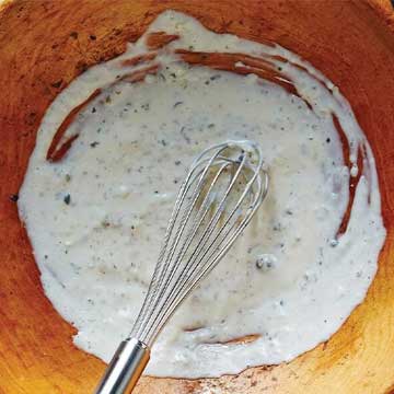 Brown bowl with Caesar salad dressing being made with a whisk off to one side. 