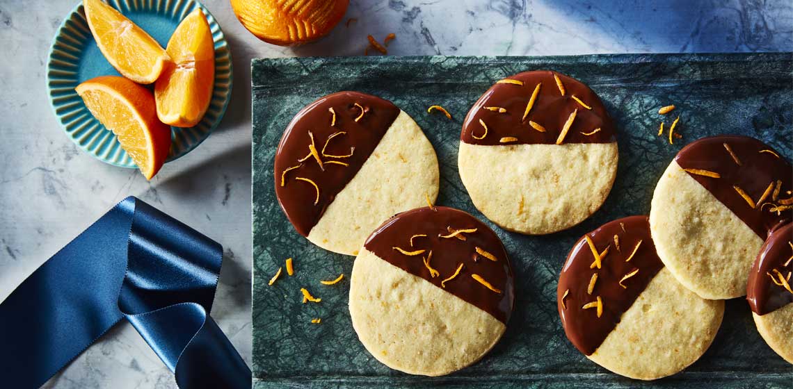 Several chocolate-clementine icebox sugar cookies half dipped in chocolate with orange zest sitting on blue plates.