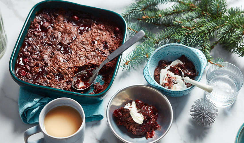 Blue baking dish with a chocolate-cranberry dump caked baked into it next to a place with a scoop of the cake and whipped cream.