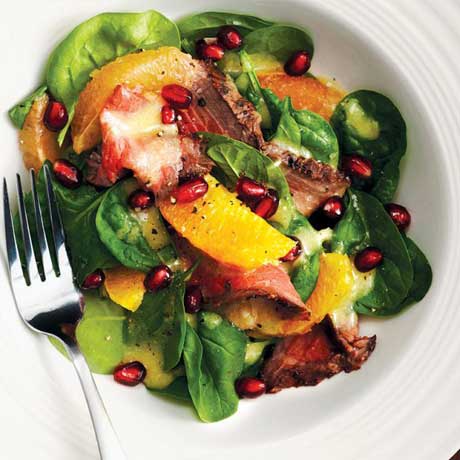 Slices of cooked beef, greens, citrus slices and pomegranate arils in a white salad bowl with a fork resting to the side.