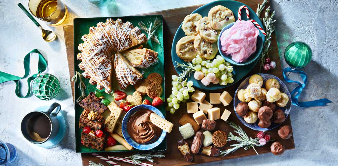 A variety of desserts including an almond kringle, chocolates, cookies, fruit cakes, candy floss and more on a several blue plates sitting on a wooden charcuterie board.