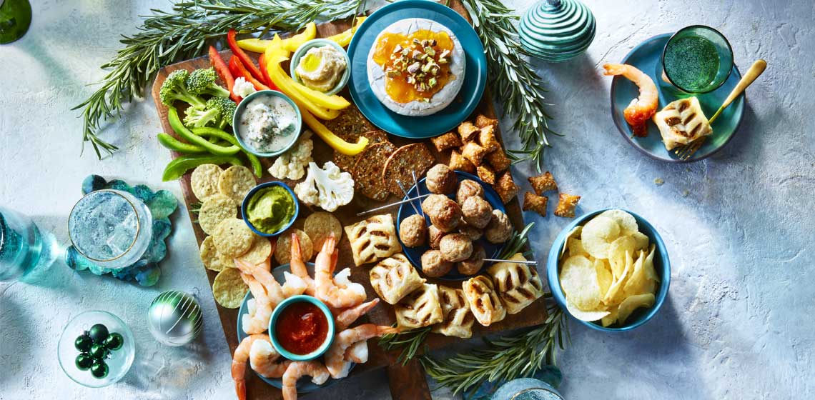 A long wooden paddle board covered with baked brie, nuts, bowl of shrimp, cheese, chips, chutney and baked appetizer pastry parcels.