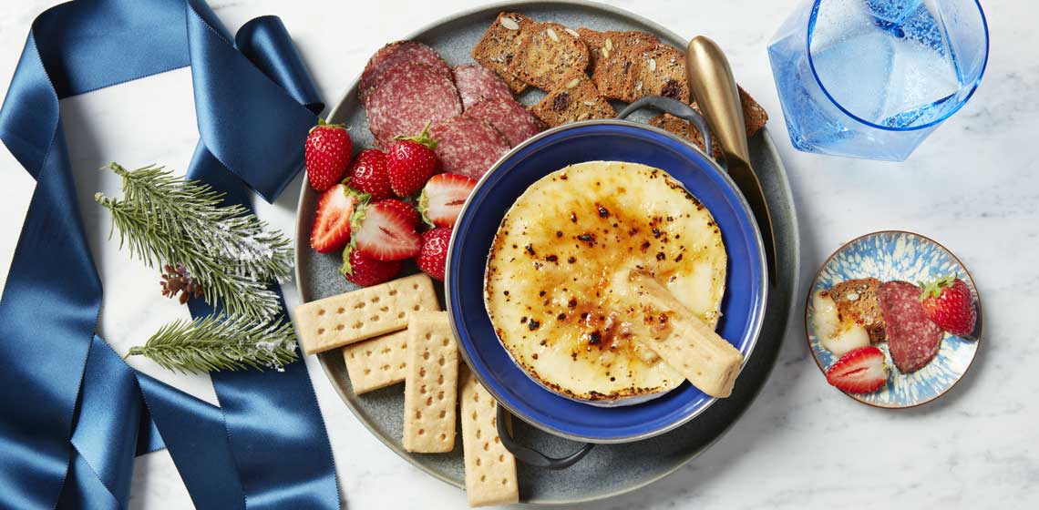 Brie with a brûléed top on a blue plate sitting next to shortbread fingers, fresh strawberries, crackers and salami meat slices.