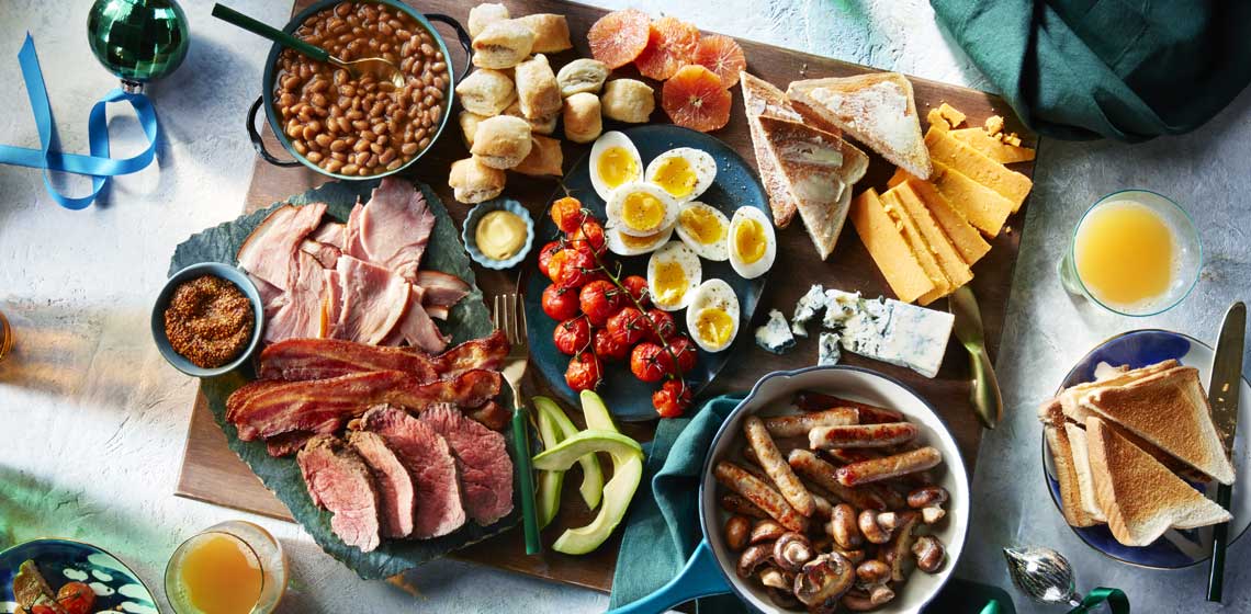 Blue platters and cast iron skillets on top of a wooden charcuterie board filled with sausages, mushrooms, soft boiled eggs, ham, rashers, toast and jam. 