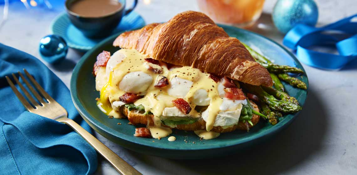 A toasted croissant filled with poached eggs, cheeses, spinach, crumbled bacon and hollandaise sauce sitting on a blue plate. 