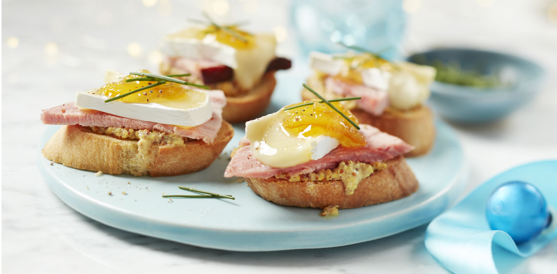 Baguette rounds topped with grainy Dijon mustard, honey smoked ham, orange marmalade and brie on a light blue plate.