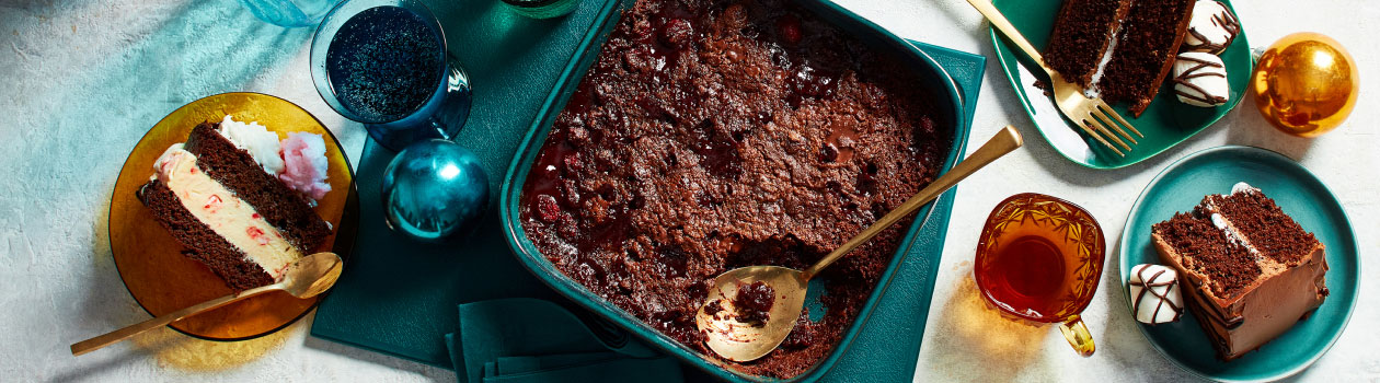 A baking pan full of dump cake, slices of other chocolate cakes on blue plates with forks next to them, a cup of tea and holiday baubles.