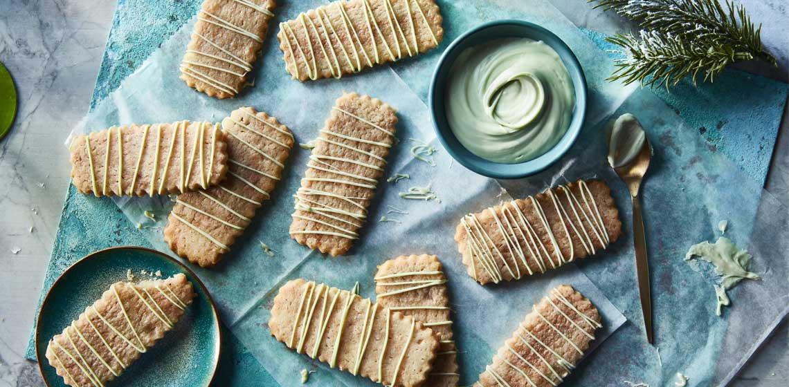 Several icebox sugar cookies drizzled with a light green melted mint flavoured baking chip on a blue plate next a blue bowl filled with the melted chips.