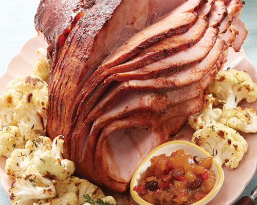 sliced ham on pink plate with small bowl of pineapple chutney surrounded by vegetables.