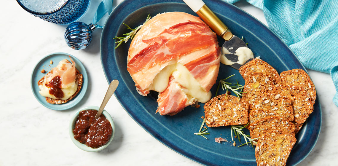 A prosciutto-wrapped wheel of brie on a blue plate next to a smattering of fresh rosemary and cracker crisps.