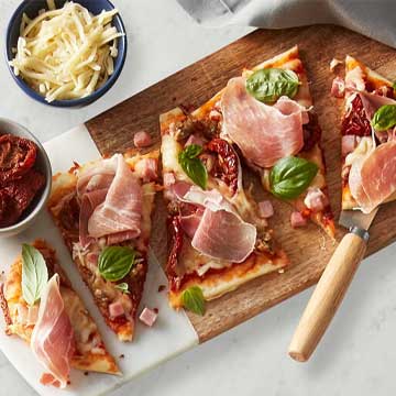 Grilled naan bread topped pizzas with sundried tomatoes, prosciutto and basil leaves on a white speckled plate and marble and wood cutting board. 