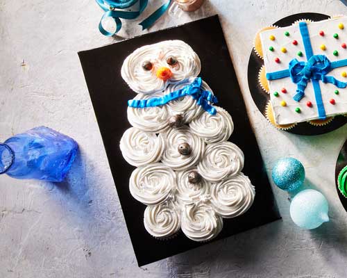 White iced cupcakes form the shape of a jolly snowman featuring a blue scarf, eyes and buttons made out of candy.