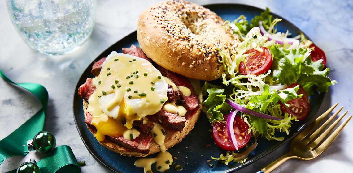 A NY-style everything bagel topped with sliced steak, poached eggs, hollandaise and chives on a blue plate.