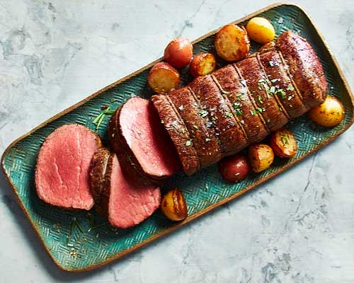 Top-down view of table with serving dish of beef wellington with roasted potatoes on a green platter.