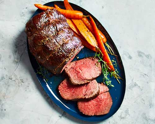 Top-down view of roast sirloin and slices of roast sirloin on dark blue serving dish with side of roasted carrots.