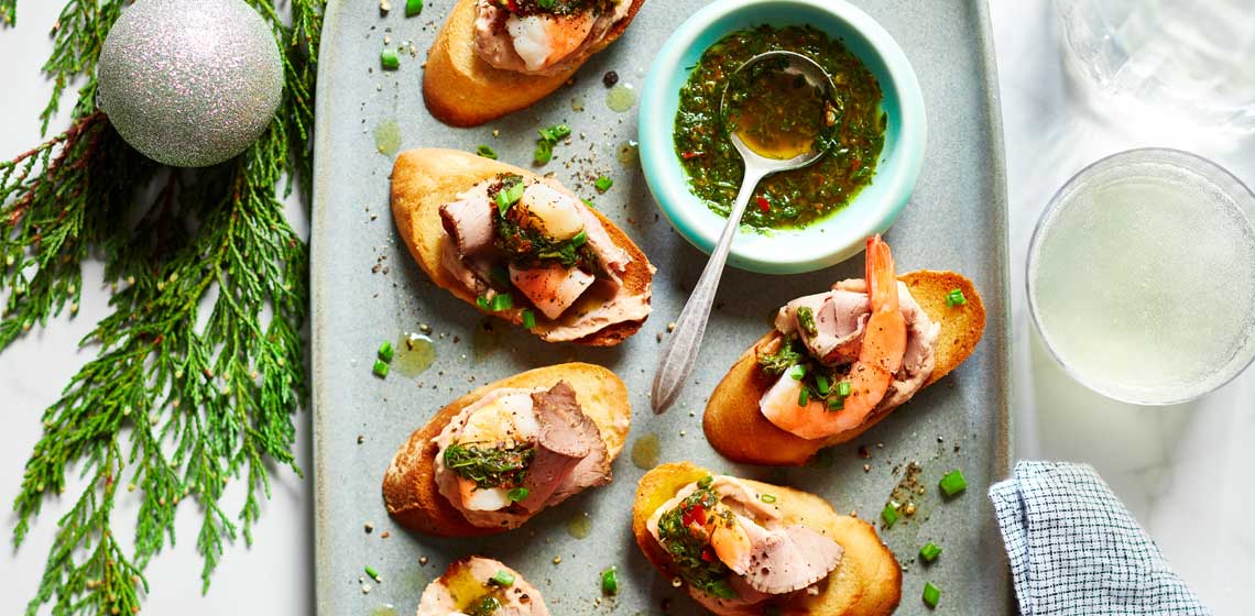 Crostini with shrimp ring shrimp and thin slices of roast beef with cocktail sauce, chimichurri and fresh herbs.