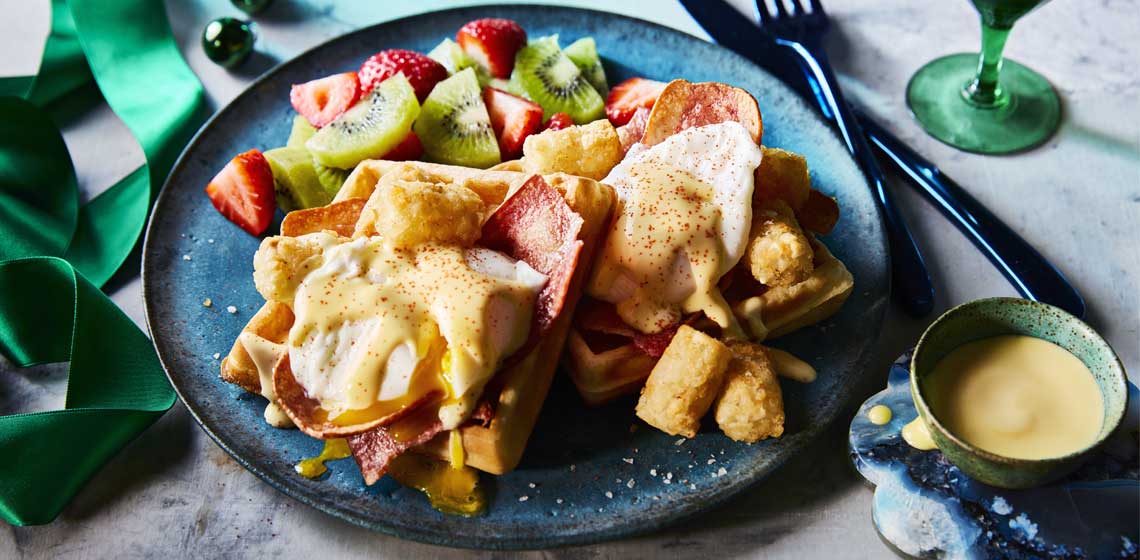 Toasted waffle topped with poached eggs, crisp turkey bacon, hollandaise sauce and potato nuggets with a side of fruit salad on a blue plate.