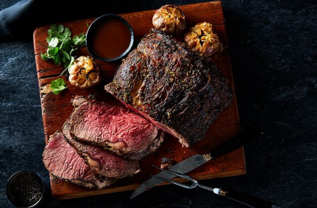 Roasted Sterling Silver prime rib on a wooden cutting board next to a roasted head of garlic with a carving knife.