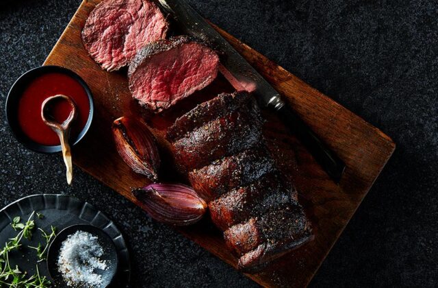 A whole roasted Sterling Silver beef tenderloin on a cutting board, with a few slices cut next to a small bowl of sauce with a wooden spoon.