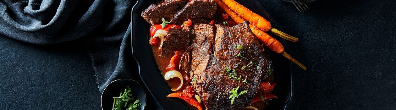 Slow-braised Tuscan sirloin roast in a black serving dish with carrots, onions and red pepper slices.