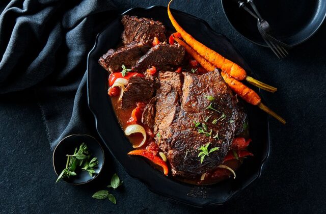 Slow-braised Tuscan sirloin roast in a black serving dish with carrots, onions and red pepper slices.