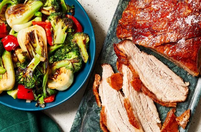 Slices of crispy pork belly on a gray platter with a bowl of charred bok choy and broccoli beside it in a blue bowl on a white countertop.