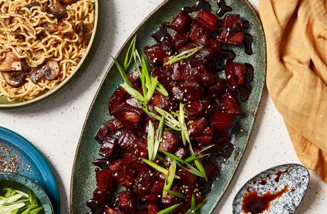 Mahogany lacquered pork belly cubes on a dark sage green oval serving platter next to a bowl of ramen noodles with mushrooms.